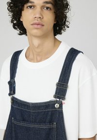 Salopette en denim avec des bretelles réglables et des accessoires en métal, dotée d'une poche avant et d'un t-shirt blanc en dessous. Couleur bleu foncé.
