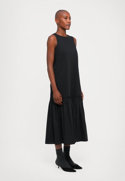 Schwarzes ärmelloses Kleid mit einem gerafften unteren Saum, das an der Oberseite glatt und am Boden fließend ist, kombiniert mit grauen Socken und schwarzen Schuhen.