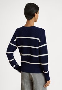 Tmavomodrý pletený sveter s bielymi vodorovnými pruhmi, žíhanou textúrou, okrúhlym výstrihom a dlhými rukávmi. Pohľad zozadu ukazuje voľnejší strih.