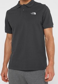 Polo gris foncé en coton, doté d'une patte de boutonnage à deux boutons et d'un petit logo blanc sur la poitrine. Coupe classique avec manches courtes.