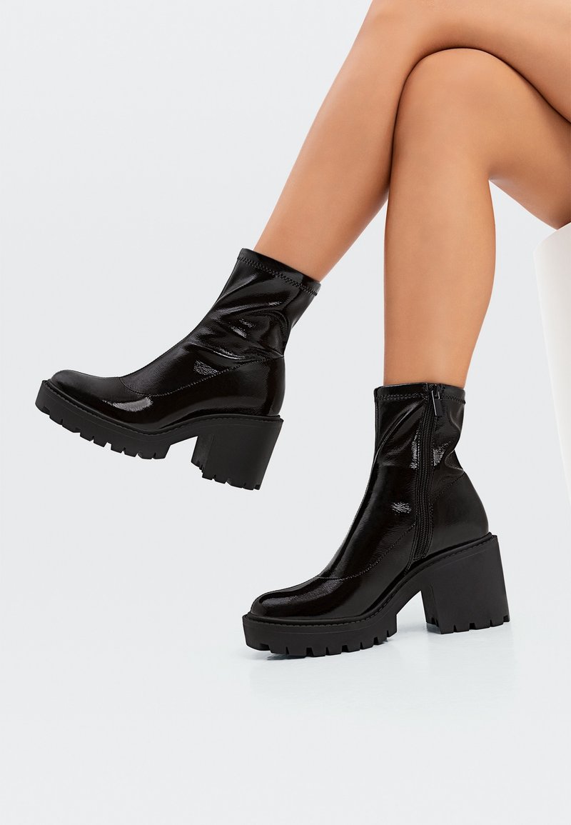 Jambes croisées, portant des bottines noires brillantes avec talons blocs et semelles crantées contrastant avec un fond clair uni.