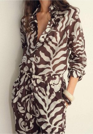 Femme portant un chemisier à motifs marron et blanc assorti à un pantalon, manches retroussées et bracelets à un poignet.