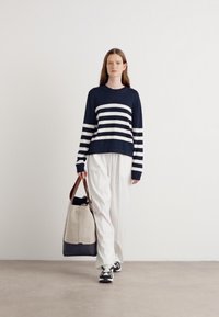 Sweater listrado em azul-marinho e branco com mangas longas, combinado com calças brancas largas e uma grande bolsa tote em bege e preto. Ténis pretos completam o visual.