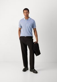 Camisa polo de color azul claro con cuello, pantalones negros y zapatos negros. Sostiene una chaqueta negra. Diseño simple con un pequeño logo en el pecho.