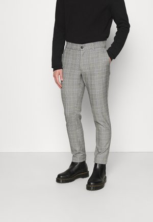 Uomo che indossa pantaloni aderenti a quadri grigi, dolcevita nero e stivali in pelle nera, in piedi con una mano in tasca.