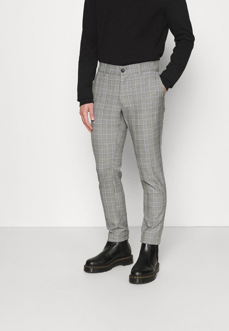 Homme portant un pantalon slim gris à carreaux, un col roulé noir et des bottes en cuir noires, debout avec une main dans la poche.