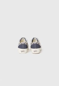 Zapatillas de lona en azul marino y crema con parte superior texturizada, diseño sin cordones y suela de goma blanca que incluye el logo de la marca en el talón.