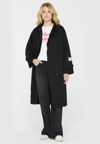 Blonde Frau trägt einen langen schwarzen Mantel über einem weißen Grafik-T-Shirt, schwarzen weiter geschnittenen Jeans und weißen Sneakers und steht mit einer Hand in der Tasche.