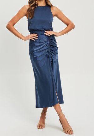 Marineblå satin kjole med en tætsiddende krop, detaljerede sider med rynker og et lårhøjt slid. Kombineret med nøgne stiletter med stropper.