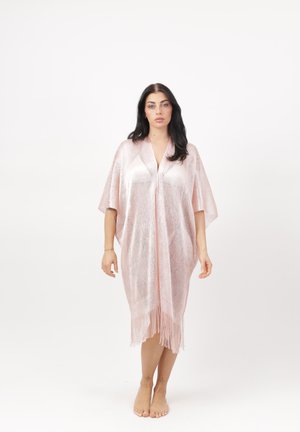 Vrouw staat blootsvoets in een glinsterende lichtroze kimono tot op de knie met wijde mouwen en een franjerand, tegen een witte achtergrond.