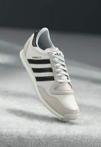 Hvit og beige treningssko med svarte striper, laget av skinn og semsket skinn, med snøring og gullfargede logoaksenter.