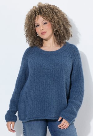 Kvinde med krøllet hår iført en blå tekstureret striktrøje og denimjeans, stående mod en lys baggrund.