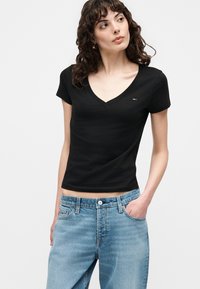 Γυναίκα με σγουρά μαλλιά που φοράει μαύρο T-shirt με V-λαιμόκοψη και ανοιχτό μπλε τζιν, ποζάροντας με το ένα χέρι στην τσέπη σε λευκό φόντο.