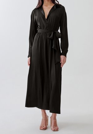 Woman wearing a long black satin shirt dress with a waist tie and nude strappy high-heeled sandals, standing against a plain background.