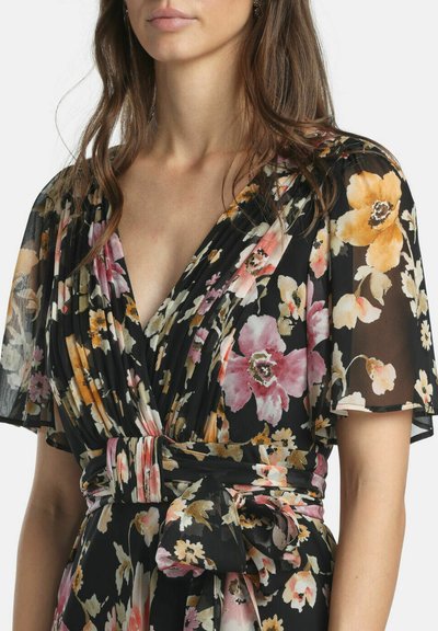 Woman wearing a black floral wrap dress with pink, orange, and yellow flowers and sheer flutter sleeves.