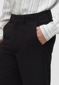 Persona che indossa pantaloni neri con la mano in tasca e una camicia bianca con righe verticali grigie.