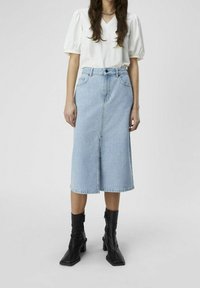 Mujer vestida con una blusa blanca de mangas abullonadas, una falda midi de mezclilla azul claro con una abertura frontal y botas de tacón negras hasta el tobillo, de pie contra un fondo liso.