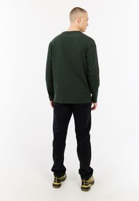 Donkergroene lange mouwen shirt van katoen, gecombineerd met zwarte denim broek en gele accenten op de schoenen, gezien van achteren.