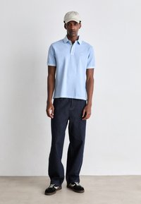 Polo shirt azul claro com mangas curtas e colarinho, combinado com calças de ganga escuras. O modelo usa um boné bege e ténis pretos.