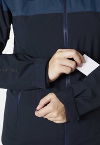 Námořnická modrá bunda se zipem, s kontrastním modrým panelem nahoře. Ruka vkládající kartu do vnitřní kapsy na zápěstí.