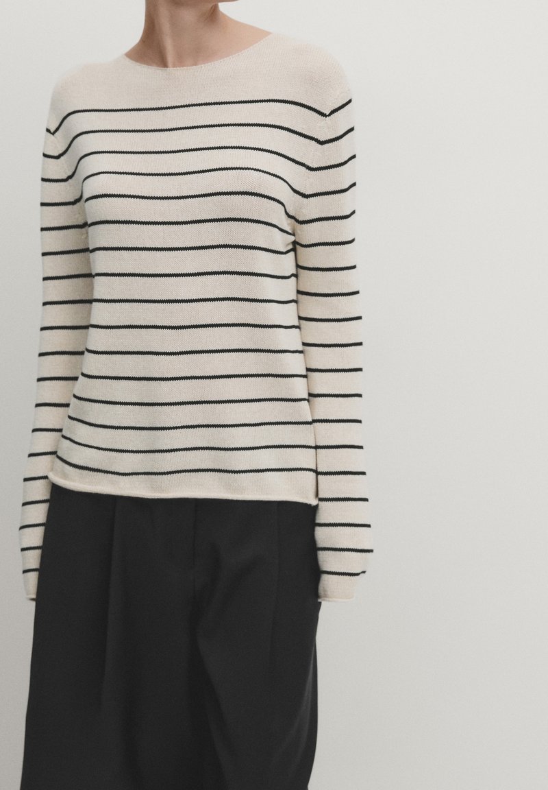 Maglione beige con strisce orizzontali nere, scollatura tonda e maniche lunghe. Il tessuto appare morbido e lavorato a maglia. Abbinato a pantaloni neri.