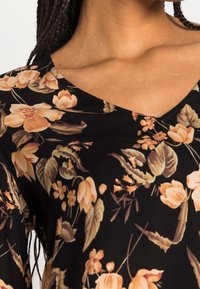Blusa nera a fiori con maniche lunghe, caratterizzata da fiori pesco e crema e foglie verdi. Design a V con una texture liscia.