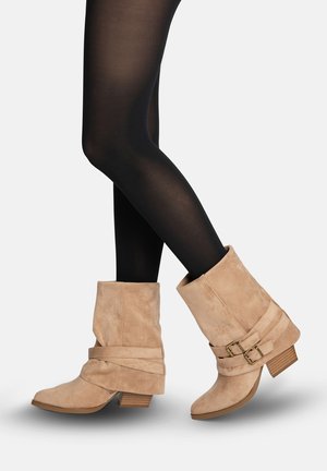 Gambe di donna con collant neri che indossa stivaletti beige scamosciati e morbidi con tacchi bassi in legno e cinturini decorativi con fibbie.