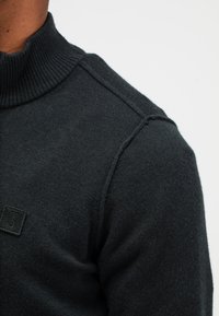 Close-up van een persoon die een donkergrijze coltrui draagt met een patch-logo op de borst tegen een witte achtergrond.