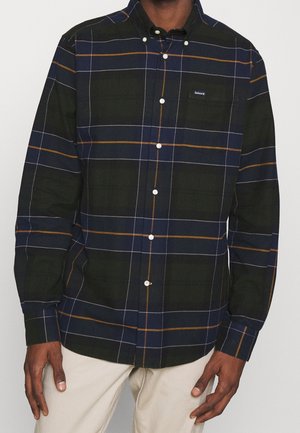 Homme portant une chemise à carreaux bleu foncé et vert avec de fines lignes blanches et orange, associée à un pantalon beige clair.