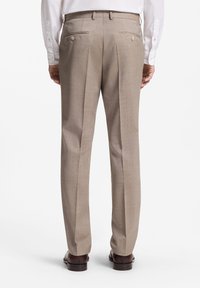 Homme portant un pantalon beige, une chemise blanche et des chaussures marron, vu de dos sur fond blanc.
