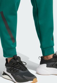 Πράσινοι αθλητικοί παντελόνες με μαύρες λεπτομέρειες στο πλάι και sneakers σε μαύρο, λευκό και καφέ χρώμα με σόλες από καουτσούκ, που διαθέτουν πλέγμα και υφή.