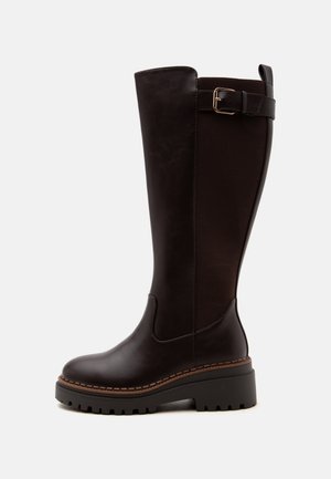 Botte haute en cuir marron foncé avec semelle crantée, panneau latéral élastique et bride décorative avec boucle près du haut sur un fond gris uni.
