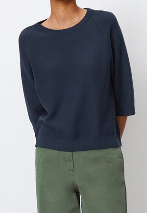 Person iført en marineblå strikket top med trekvartærmer og olivengrønne bukser, stående foran en ensfarvet hvid baggrund.