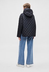 Čierna prešívaná bunda s kapucňou a diamantovým vzorom, v kombinácii so širokými modrými rifľami a bielymi teniskami. Zobrazený pohľad zozadu.