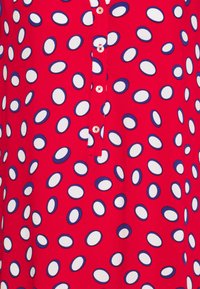 Červená košile se zapínáním na knoflíky, zdobená abstraktním vzorem bílých a námořnických modrých oválných tvarů. Hladká textura látky.