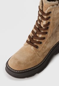 Stivaletto alla caviglia in suede tan con lacci marroni, suola in gomma e dettagli di cucitura. Presenta una fodera morbida e texture per un comfort aggiuntivo.