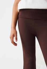 Leggings marron extensibles avec une taille haute, une texture lisse et une coupe ajustée, accompagnés d'une main posée à proximité.