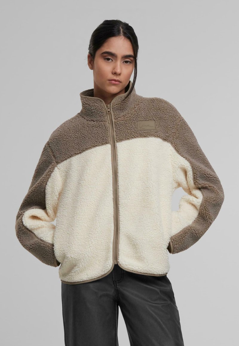Chaqueta de forro polar en color crema y marrón, con cuello alto y cierre de cremallera. Destacada por su superficie texturizada y un pequeño parche con el logo.