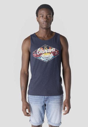 Joven con camiseta sin mangas azul marino con texto "Okinawa Beach" y diseño colorido, combinado con shorts de mezclilla azul claro remangados.