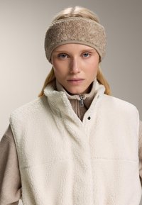 Beige fleecová čelenka s měkkým, texturovaným povrchem, spárovaná s krémově bílou, nadýchanou vestou a hnědým topem s vysokým límcem, který má zip.