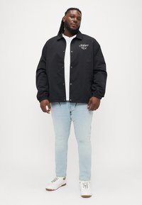 Crna podstavljena jakna s dugmadi na preklop, podesivim manžetama i bijelim logotipom na prsima, u kombinaciji sa svijetloplavim farmerkama i bijelim tenisicama.