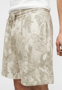 Shorts beiges avec un motif discret de feuilles, taille à cordon, poches latérales, portés par une personne avec une chemise blanche visible à la taille.