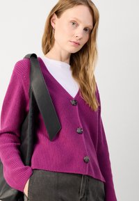 Purple knit cardigan with a V-neck, three black buttons, and ribbed texture, worn over a white T-shirt, with a black shoulder bag.
