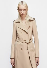 Bežový dvojradový trenčkot s opaskom v páse, epauletmi na pleciach, hladkým materiálom a gombíkovými detailami. Klasický golier a dlhé rukávy.