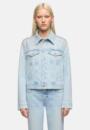 Donna con capelli biondi lunghi che indossa una giacca di jeans azzurro chiaro e jeans, giacca decorata con piccoli borchie d'argento sulla metà superiore.