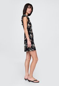 Čierne kvetinové šaty s volánovými rukávmi, s priliehavým živôtikom a rozšírenou sukňou. Vzor zahŕňa malé farebné kvety. Nosené s čiernymi sandálmi.