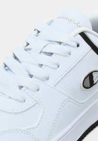 Λευκό αθλητικό παπούτσι με λείο συνθετικό δέρμα, διακοσμητικά πλεκτά μοτίβων, επίπεδα κορδόνια και ετικέτα με το λογότυπο με ασημί λεπτομέρειες στη γλώσσα.