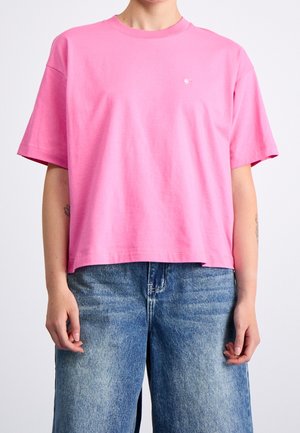 Person wearing a loose, short-sleeve pink t-shirt paired with faded blue denim jeans, standing against a plain background.