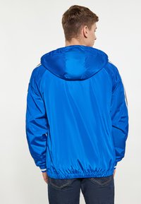 Giacca in nylon blu con cappuccio, orlo elastico e accenti a righe sulle maniche, cuciture visibili e texture liscia, indossata sopra jeans scuri.