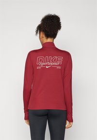 Crvena majica dugih rukava za sport s visokim ovratnikom, s bijelim logotipom "Nike Sportswear" na leđima. Meka, strukturirana tkanina, uski kroj.